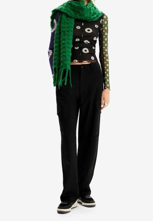 Femme portant un crop top à manches longues à motifs multiples, un pantalon cargo noir, des baskets noires et une écharpe texturée vert vif.