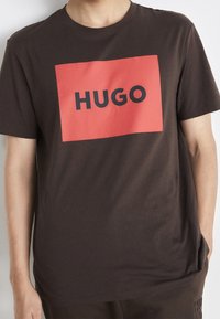 Hnědé bavlněné tričko s velkým červeným obdélníkovým motivem s nápisem „HUGO“ tučnými černými písmeny. Krátké rukávy, volný střih.