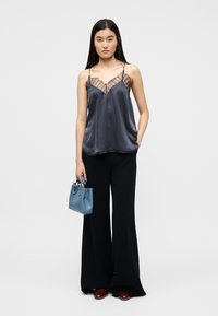 Femme portant un caraco en satin gris foncé transparent, un pantalon noir à jambes larges, des chaussures bordeaux, et tenant un petit sac à main bleu.