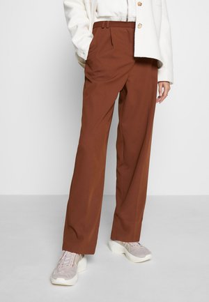 Femme portant un pantalon large marron taille haute, une veste crème, et des baskets rose clair, debout avec une main dans la poche devant un fond blanc.