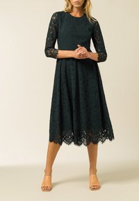 Kanten jurk in donkergroen met driekwart mouwen en een uitlopende rok. Bevat een gegolfde zoom en intrikate kantdetails.