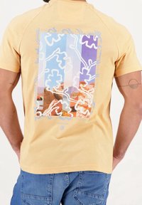 Kortærmet gul t-shirt med et farverigt grafik på ryggen, der viser abstrakte mønstre og landskabsbilleder. 100% bomuldsstof, almindelig pasform.