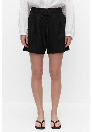 Vrouw draagt zwarte shorts met hoge taille en strikceintuur, witte blouse met lange mouwen en zwarte teenslippers, staand tegen een witte achtergrond.
