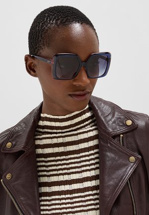 Persona con capelli corti che indossa grandi occhiali da sole quadrati, un top a righe a coste e una giacca in pelle marrone scuro con bottoni dorati.