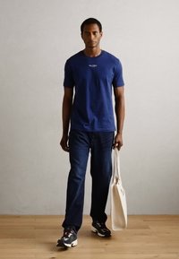 Marineblå t-skjorte, denim jeans og mønstrede sneakers; modellen holder en lys beige tote bag; står på et tregulv foran en ensfarget vegg.