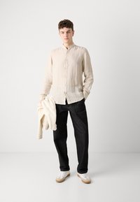 Camicia di lino button-up di un leggero beige, jeans neri a gamba dritta e sneakers bianche con suola in gomma, mentre tiene una giacca beige.