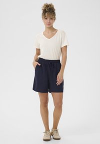 Svetlo béžové tričko s výstrihom do V skombinované s tmavomodrými šortkami na šnúrku. Šortky majú vrecká a voľný strih. Outfit dopĺňajú svetlé topánky.