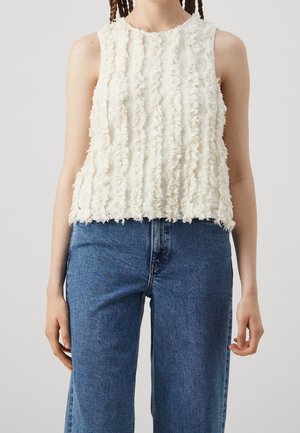 Haut blanc texturé sans manches avec des bandes à franges verticales porté avec un jean en denim bleu taille haute sur une personne aux cheveux tressés.