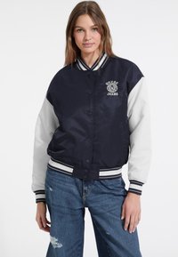 Marineblauwe en witte bomberjack met een satijnen afwerking, geribbelde kraag en drukknopen aan de voorkant. Bevat het "Guess Jeans" logo op de borst.