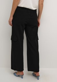 Zwarte cargo broek met een losse pasvorm, elastische tailleband, zijzakken en een gladde textuur. Draagt met blauwe hakken mules.