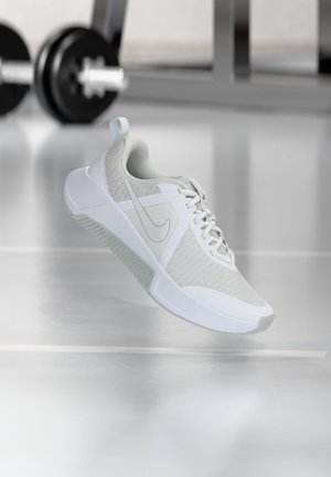 Zapatilla deportiva blanca con parte superior de malla y material sintético, logo visible de Nike, flotando sobre un suelo de gimnasio brillante con equipo de levantamiento de pesas desenfocado en el fondo.