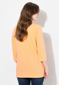 Oranges T-Shirt mit Drei-Viertel-Ärmeln, lockerer Passform, weichem Material und abgerundetem Saum, kombiniert mit dunkelblauen Jeans.