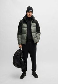 Puffer jacket in olive green and black, paired with a black hoodie and sweatpants. Black beanie and backpack with a water bottle visible.