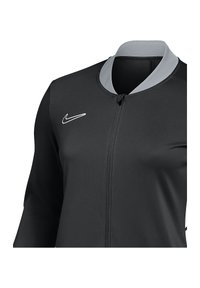 Schwarze Jacke mit einem grauen Kragen, mit einem Frontreißverschluss, gefertigt aus glattem Stoff. Nike-Logo in Weiß auf der linken Seite. Lange Ärmeln.