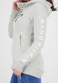 Jasnoszara bluza z kapturem ze sznurkami, dużą boczną kieszenią i nadrukowanym tekstem na rękawie. Na piersi znajduje się małe logo w kształcie serca.