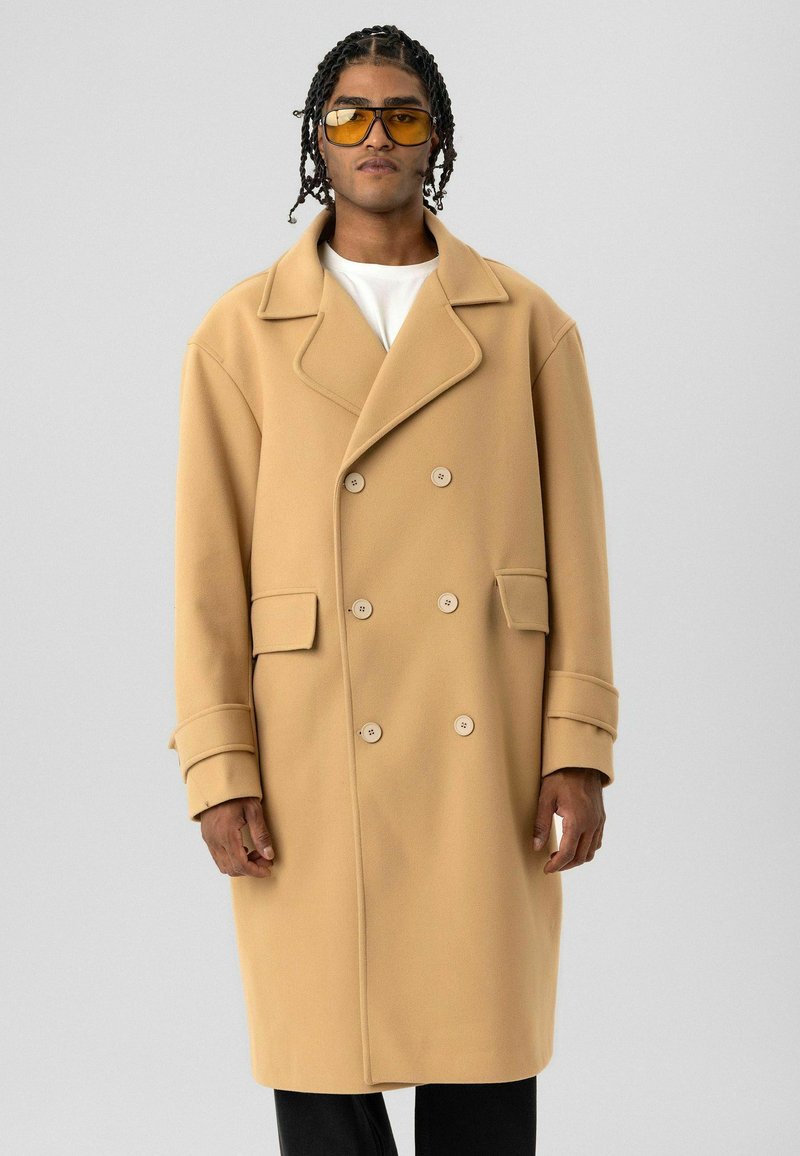 Beige dubbelgeknoopte jas met brede revers, knoopmanchetten, voorzakken en een gladde textuur. Draagt over een wit overhemd.