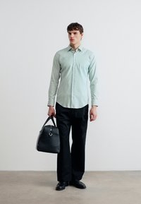 Άνδρας με ανοιχτό πράσινο πουκάμισο με κουμπιά και μαύρο παντελόνι, κρατώντας μαύρη δερμάτινη τσάντα στο δεξί χέρι, στέκεται μπροστά σε απλό λευκό τοίχο.