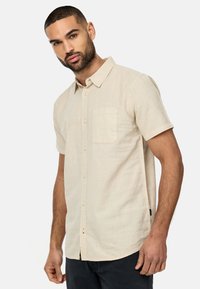 Beige kortærmet skjorte med knapper og brystlomme, letvægtsstof og en subtil tekstureret overflade. Kurvet bund med sidesømme.