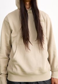 Sudadera beige con un bolsillo delantero, puños y dobladillo acanalados, y capucha con cordón. Textura suave con detalles de marca mínimos.