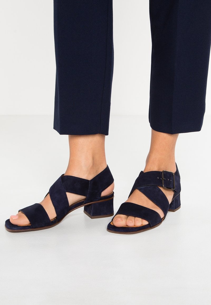 Pieds portant des sandales à talon bloc en daim bleu foncé avec des brides croisées et une boucle à la cheville, associées à un pantalon court foncé.