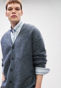 Young man wearing a blue fuzzy cardigan over a light blue striped button-up shirt with rolled-up sleeves against a plain background.