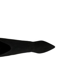 Bottine en daim noir avec un bout pointu, une silhouette effilée et une texture lisse, présentant un détail en V sur les côtés.