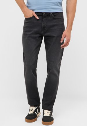Mann trägt schwarze Slim-Fit-Jeans, hellblaues Hemd und schwarze Sneakers mit weißen Schnürsenkeln und Gummisohlen, steht mit einer Hand in der Hosentasche.