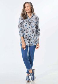 Himmelblaues, blumiges Hemd mit Knöpfen und langen Ärmeln, kombiniert mit hochgekrempelten blauen Jeans und blauen gemusterten Sneakers. Glattes Material mit auffälligen Mustern.