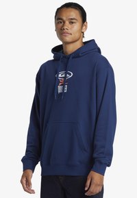 Sudadera azul marino con un bolsillo tipo canguro, con un logo gráfico en blanco y naranja en el pecho. Hecha de una tela suave y texturizada.