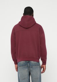 Felpa oversize borgogna con un ampio cappuccio, polsini e orlo a costine. Presenta una texture morbida e spalle scese, abbinata a jeans azzurri chiari.