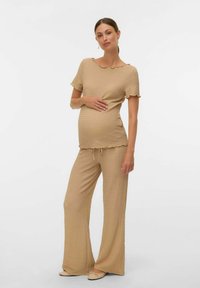 Donna incinta in cima a maniche corte beige con texture e pantaloni larghi abbinati, in piedi con una mano sul pancione su uno sfondo bianco.