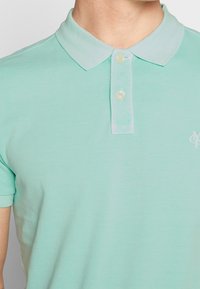 Jasnozielona koszulka polo z kołnierzykiem, zapinana na dwa guziki, z subtelnym logo na piersi. Gładka, lekko teksturowana tkanina.