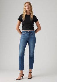 Camiseta negra cortada, diseño ajustado. Jeans rectos azules con un ligero desvanecimiento. Tacones negros de tiras. Fondo gris neutro.