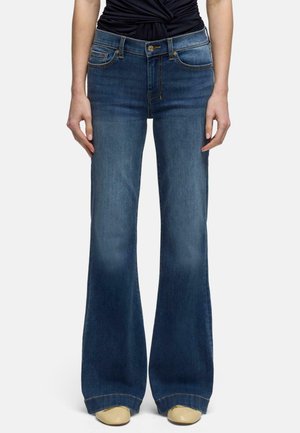 Person iført højtaljede, mørkeblå vide jeans med knap og lynlås foran, kombineret med beige sko og en sort top med rynkede detaljer.