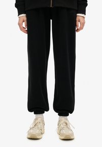 Pantalones de chándal negros con un ajuste relajado, puños acanalados en los tobillos y un detalle de logo bordado. Combinados con zapatillas de color claro.