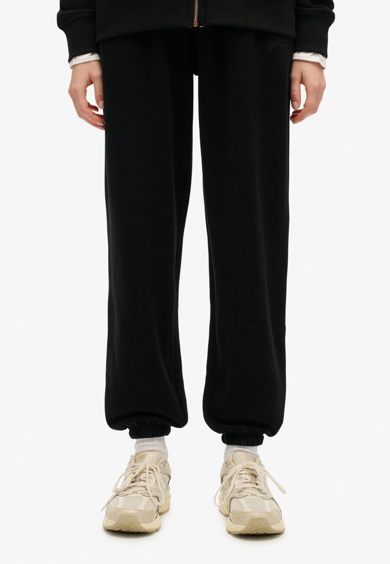 Pantalones de chándal negros con un ajuste relajado, puños acanalados en los tobillos y un detalle de logo bordado. Combinados con zapatillas de color claro.