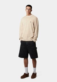 Sudadera beige de cuello redondo, shorts de mezclilla negros, zapatos marrones, calcetines blancos. Características destacadas: logo en la sudadera, bolsillos en los shorts, corte relajado.