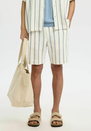 Person som har på seg kremfargede og blå stripete shorts og matchende kortermet skjorte, holder en beige stoffveske og har på seg beige sandaler.