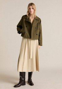 Olive green cropped jacket with double-breasted black buttons over a beige pleated dress, accompanied by knee-high black boots.