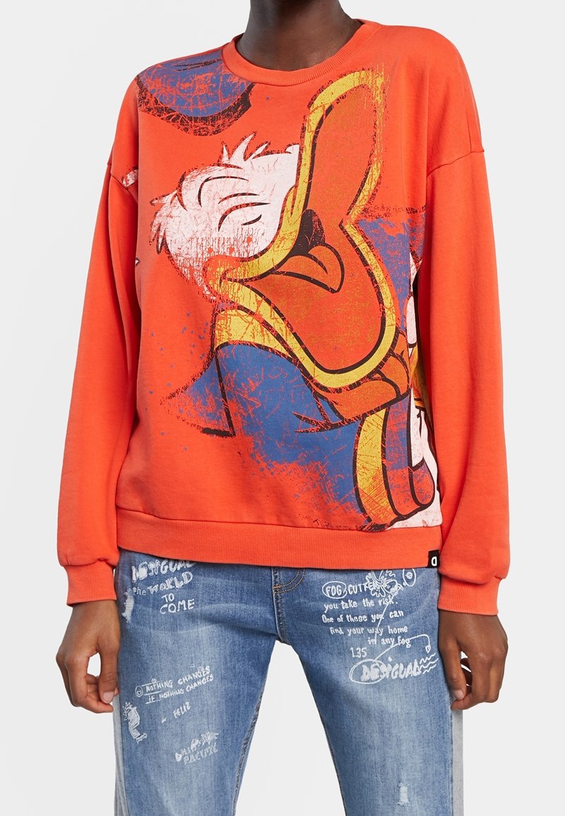 Personne portant un sweat-shirt orange vif avec un grand graphisme usé du visage de Donald Duck et un jean bleu avec du texte et des dessins imprimés en blanc.