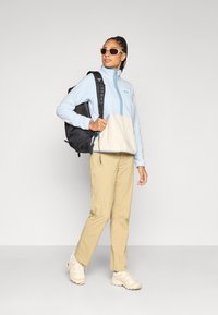 Ανοιχτό μπλε φλις πουλόβερ με κρεμ κάτω μέρος, μπεζ παντελόνια και λευκά αθλητικά παπούτσια. Μαύρο σακίδιο πλάτης, με αξεσουάρ γυαλιά ηλίου.