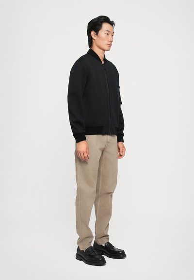 Sort bomberjakke med ribbede manchetter og krave, parret med lysebeige bukser. Modellen bærer sorte snørebåndssko. Simpelt, rent design.