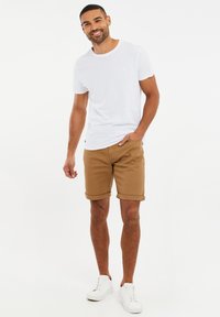 Camiseta blanca de algodón con escote redondo, combinada con pantalones cortos marrones plegados y zapatillas blancas, sobre un fondo liso.