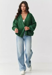 Zielony, puszysty kardigan z głębokim dekoltem w kształcie litery V, szerokimi rękawami i luźnym krojem, w połączeniu z jasnoniebieskimi dżinsami o prostym kroju i białymi trampkami.