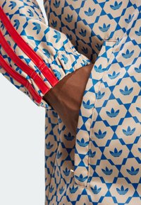 Chaqueta con patrón que presenta una tela beige con formas geométricas azules y los icónicos logos de trébol. Las mangas tienen franjas rojas que las acentúan.