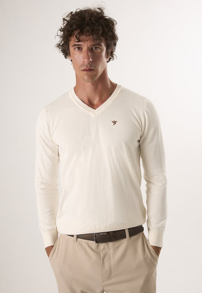 Pull en V crème en tissu doux, doté de manches longues et d'un petit logo de couleur brune représentant un oiseau sur la partie gauche de la poitrine, associé à un pantalon beige.