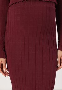 Burgundské žebrované šaty s přiléhavým střihem, dlouhými rukávy a strukturovaným materiálem s mírným elastickým efektem. Vyřezávaný okraj na horní části.