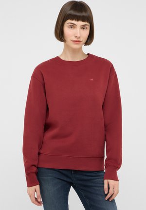 Junge Frau mit kurzen braunen Haaren, die einen schlichten roten Pullover und dunkelblaue Jeans trägt und vor einem einfarbigen hellen Hintergrund steht.