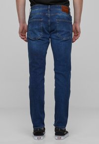 Jean en denim avec un délavage bleu moyen, coupe droite, cinq poches et une patch en cuir marron à l'arrière de la taille. Chaussures à semelle en caoutchouc visibles.