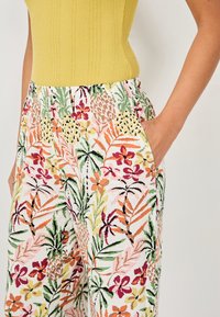 Pantalones con estampado floral sobre una base blanca, que presentan vibrantes diseños botánicos en verde, rosa, naranja y amarillo. Incluyen bolsillos laterales.